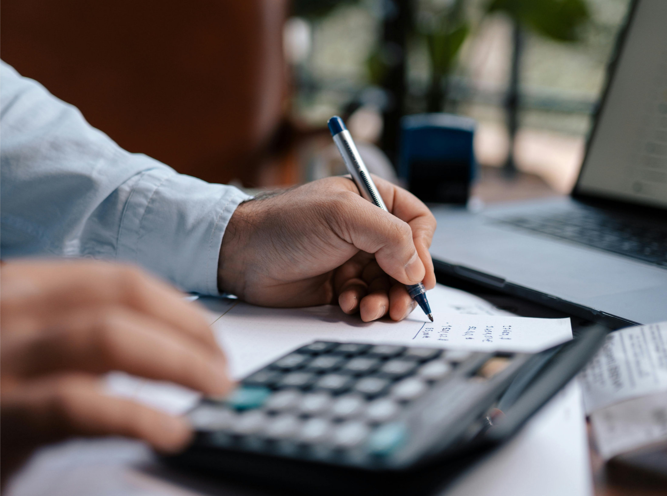 Mãos de um homem em que uma segura uma caneta sobre papel e outra digita em calculadora com computador portátil ao fundo